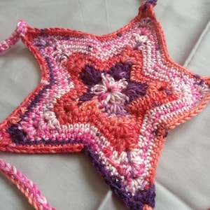 May include: A star-shaped crochet item in pink, orange, and purple hues. It features a central floral motif and braided ties. The item is likely a decorative piece or accessory, possibly a handmade item.