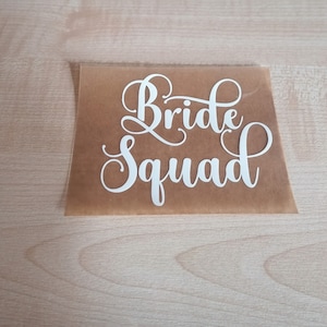 Puede incluir: Calcomanía de vinilo blanco con el texto "Bride Squad" en una fuente cursiva sobre un fondo de papel marrón.