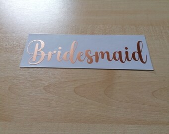 May include: A rectangular sticker with the word "Bridesmaid" in elegant, rose gold script. The sticker is on a white background, set against a light wood surface. Ideal for wedding decorations.
