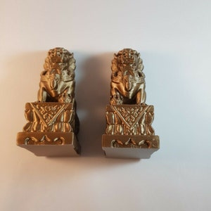 May include: Two gold-coloured Foo Dog statues. Each statue features a detailed lion-like figure atop a decorative base with intricate carvings. The statues are displayed on a white surface.