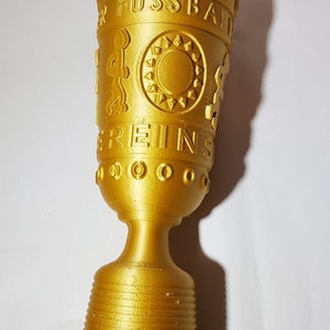 May include: A gold-colored trophy with the words "DFB Pokal" and "Vereins" embossed on the cup. The trophy has a detailed design with a sunburst and a figure of a soccer player.