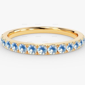 Puede incluir: Un anillo de banda de oro con una fila de piedras preciosas de color azul claro. El anillo presenta una banda delgada y las piedras preciosas están espaciadas uniformemente. El anillo está sobre un fondo blanco.