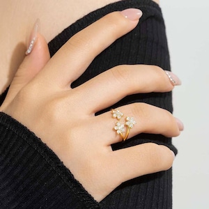 Puede incluir: Un anillo dorado con tres diseños en forma de flor, cada uno adornado con pequeñas piedras preciosas transparentes. El anillo se lleva en un dedo, con las flores colocadas para crear una estética floral delicada. La banda del anillo es delgada y elegante.