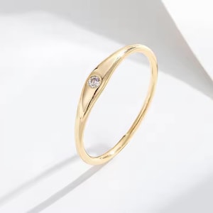 Puede incluir: Un delicado anillo de oro con una sola piedra preciosa pequeña y transparente engastada en una sección ligeramente más ancha de la banda. El anillo tiene un acabado liso y pulido y un diseño simple y elegante.