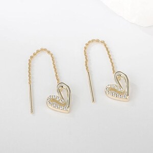 Puede incluir: Un par de pendientes dorados en forma de corazón. Cada pendiente presenta un colgante de corazón con pequeños cristales transparentes y una delicada cadena. Los pendientes se muestran sobre un fondo blanco. Joyería romántica.