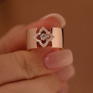 Pode incluir: Um anel de ouro rosa com uma pedra transparente em forma de diamante engastada em um design de trevo de quatro folhas. O anel é largo e tem uma banda plana.