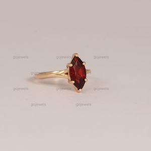 May include: A gold ring featuring a marquise-cut, deep red gemstone. The ring's band is a simple, elegant design, and the gemstone is the focal point. The ring is set against a neutral background.