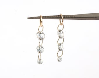 May include: A pair of gold-toned earrings with a dangling design. Each earring features a small hoop at the top, connected to a series of four clear, round gemstones. The gemstones are linked by delicate gold chains, creating a cascading effect.