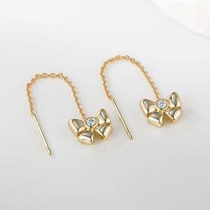 May include: A pair of gold-colored bow earrings with a small, clear gemstone in the center of each bow. The earrings have a chain that curves over the ear and a straight post. The background is a soft, off-white color.