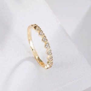 Puede incluir: Un anillo dorado con una serie de diseños en forma de corazón, cada uno adornado con pequeñas piedras brillantes. El anillo se presenta sobre un fondo blanco suave, resaltando sus delicados detalles y su elegante diseño.