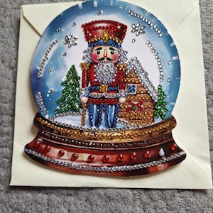 Puede incluir: Tarjeta de felicitación navideña con diseño de bola de nieve. En el interior, un soldado cascanueces, una pequeña casa y un árbol de Navidad. La bola y la base están decoradas con pedrería. La tarjeta está sobre un sobre crema.
