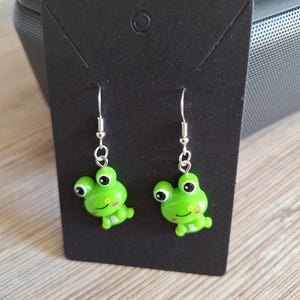 May include: A pair of green frog earrings with black eyes and pink cheeks. The earrings are made of a shiny material and hang from silver-colored hooks. The earrings are displayed on a black card.