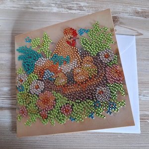 Puede incluir: Tarjeta de felicitación hecha a mano con un diseño de gallo y polluelos, adornada con pedrería de colores. La obra de arte incluye un gallo con tonos rojos, naranjas y marrones, rodeado de acentos florales verdes, azules y rosas. La tarjeta se acompaña de un sobre blanco.
