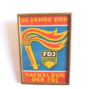 May include: A rectangular pin with a gold frame featuring a torch design with red and yellow flames. The pin has the text "30 JAHRE DDR" and "FACKELZUG DER FDJ" in red and yellow. A blue background completes the design.