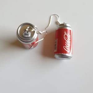 Può includere: Un paio di orecchini d'argento a forma di lattine di Coca-Cola. Le lattine sono rosse con scritte bianche e un top argentato.