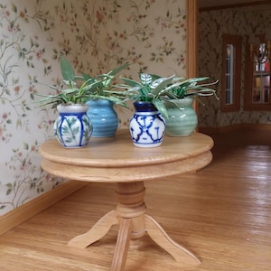 May include: A miniature wooden table with a round top and three-legged base. Four small ceramic vases with green plants sit on the table. The vases are blue, green, and white with decorative patterns. The room has floral wallpaper and hardwood floors.