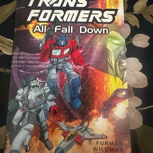 May include: A comic book titled "TRANSFORMERS All Fall Down" features a dynamic illustration of Autobots and Decepticons in action. The cover showcases Optimus Prime, a jet, and other characters against a fiery backdrop. The authors' names are also visible.