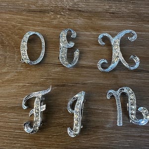 Op de afbeelding: Een verzameling zilverkleurige letterbroches, waaronder de letters O, E, X, F, I en n. Elke broche is bezet met kleine, heldere kristallen. De broches liggen op een houten oppervlak.