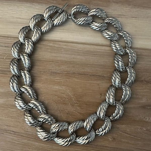May include: Silver-toned necklace featuring a series of large, textured oval links. The links are connected to form a continuous chain, with a T-bar clasp. The necklace is displayed on a wooden surface.