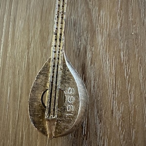Pode incluir: Um broche de mandolim prateado, em estilo vintage. O broche apresenta um design detalhado do instrumento, incluindo cordas, uma boca e o número "966" gravado no corpo. Fundo de madeira.