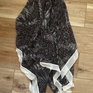 Puede incluir: Una bufanda gris oscuro y blanca con un estampado de piel de serpiente. La bufanda tiene un borde blanco y está drapeada sobre una superficie de madera. La bufanda está hecha de un material ligero.