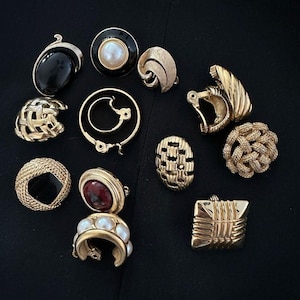 Puede incluir: Colección de pendientes vintage de clip en varios tonos dorados y diseños. Incluyen una piedra negra con ribete dorado, una perla con borde negro y patrones trenzados y geométricos. Los pendientes se muestran sobre una superficie negra.