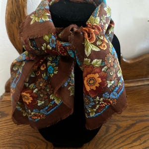 May include: A brown square scarf featuring a vibrant floral pattern with orange, yellow, light blue, and green blossoms and leaves. The fabric has a soft texture and finished edges with a delicate fringe detail along the perimeter of the scarf.