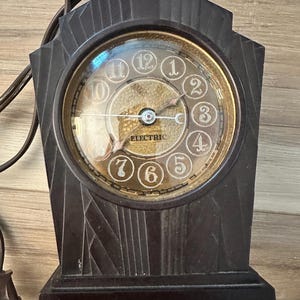 Op de afbeelding: Een donkerbruine elektrische klok met een ronde wijzerplaat en een decoratief, geometrisch ontwerp. De wijzerplaat heeft witte cijfers en wijzers, met het woord "ELECTRIC" in het midden. De klok is aangesloten op een stopcontact.