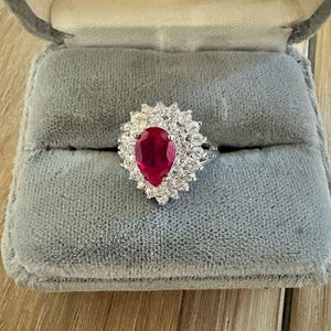 May include: A silver-toned ring featuring a large, pear-shaped, deep red center stone surrounded by a halo of numerous small, clear, round-cut stones. The ring is displayed resting inside a grey, velvet-lined jewelry box with a hinged lid.