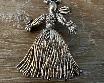 Puede incluir: Broche vintage con forma de muñeca, hecho de metal color bronce. La muñeca tiene un vestido detallado, brazos extendidos y un lazo en la cabeza. El broche está sobre una superficie de madera.