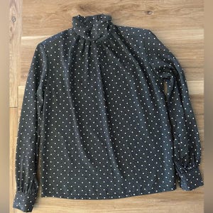 Könnte beinhalten: Eine schwarze Bluse mit weißen Punkten, hohem Kragen und langen Ärmeln.