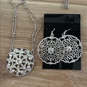 Pode incluir: Um conjunto de joias prateadas com colar e brincos. O colar tem um pingente redondo com uma pedra transparente central e pedras menores. Os brincos são circulares com um design semelhante, em um cartão preto.