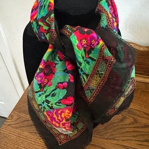 May include: A black scarf featuring a vibrant floral pattern with bright pink, orange, yellow, and green colors. The scarf has a decorative border with geometric shapes and intricate details, draped over a black display form.