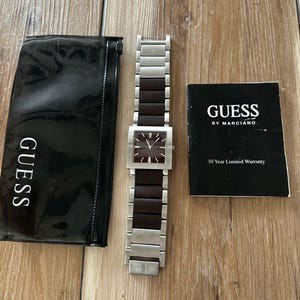 May include: A Guess watch with a square, dark face and a silver and brown strap. The watch is displayed with a black pouch and a booklet that reads "GUESS BY MARCIANO" and "10 Year Limited Warranty."
