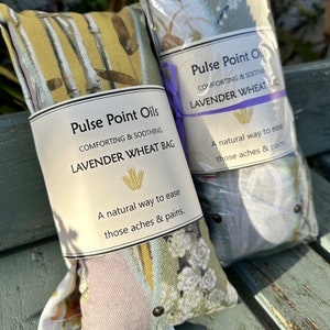 May include: Two lavender wheat bags with floral patterns and the text "Pulse Point Oils, Comforting & Soothing, Lavender Wheat Bag". The bags are wrapped in clear plastic and tied with a purple ribbon. The text "A natural way to ease those aches & pains" is also visible.