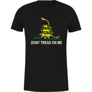 Op de afbeelding: Zwart T-shirt met een gele en zwarte opgerolde slangengrafiek en de witte tekst "DONT TREAD ON ME". De slang staat boven groen gras. Het T-shirt heeft een ronde hals en korte mouwen.