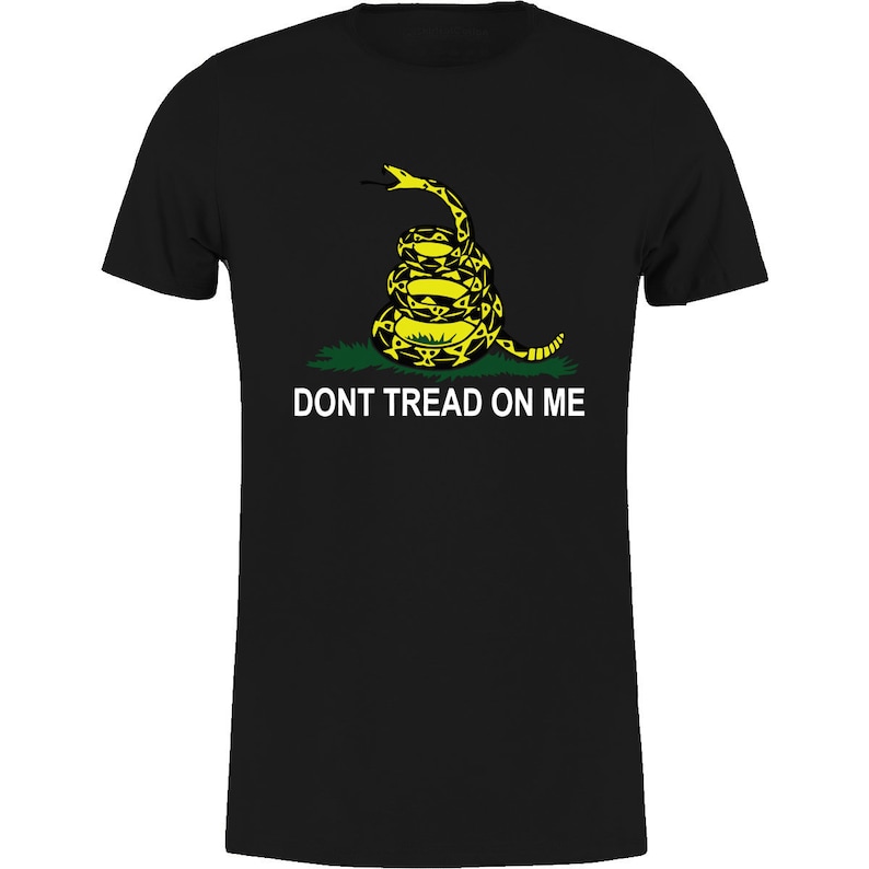 Op de afbeelding: Zwart T-shirt met een gele en zwarte opgerolde slangengrafiek en de witte tekst "DONT TREAD ON ME". De slang staat boven groen gras. Het T-shirt heeft een ronde hals en korte mouwen.