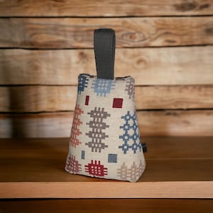 Puede incluir: Un tope de puerta de tela con forma de cu&ntilde;a, con una base de color crema y un patr&oacute;n repetitivo de peque&ntilde;os motivos geom&eacute;tricos de punto de cruz en tonos azul, rojo, marr&oacute;n y beige. Un asa de tela gris oscuro est&aacute; unida a la parte superior.