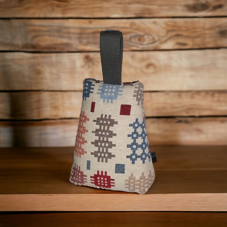 Puede incluir: Un tope de puerta de tela con forma de cu&ntilde;a, con una base de color crema y un patr&oacute;n repetitivo de peque&ntilde;os motivos geom&eacute;tricos de punto de cruz en tonos azul, rojo, marr&oacute;n y beige. Un asa de tela gris oscuro est&aacute; unida a la parte superior.
