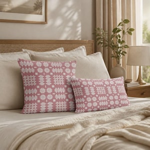 May include: Two throw pillows featuring a dusty rose pink background with a repeating white geometric pattern of small squares and cross-like shapes. One pillow is square and the other is rectangular, both displaying the same consistent design.