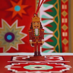 May include: A wooden Christmas ornament in the shape of a nutcracker soldier, with a red uniform and gold crown. The ornament is suspended by a red ribbon. The background features a red and patterned design.