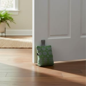Puede incluir: Un tope de puerta rectangular de tela en un tono verde vibrante con un patr&oacute;n geom&eacute;trico gris claro repetitivo. El tope incluye una peque&ntilde;a asa de tela gris en la parte superior y una etiqueta negra en el lateral, colocado junto a una puerta.
