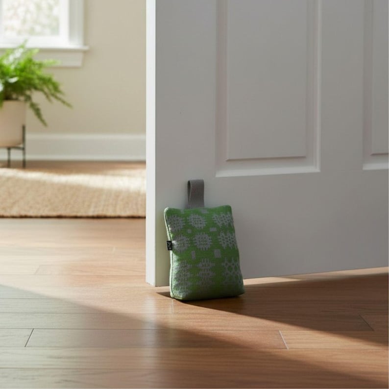 Puede incluir: Un tope de puerta rectangular de tela en un tono verde vibrante con un patr&oacute;n geom&eacute;trico gris claro repetitivo. El tope incluye una peque&ntilde;a asa de tela gris en la parte superior y una etiqueta negra en el lateral, colocado junto a una puerta.