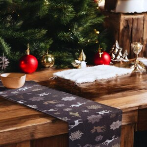May include: A dark brown table runner with a festive design featuring gold snowflakes and reindeer. The runner is laid out on a wooden table with a white fuzzy placemat and Christmas decorations.