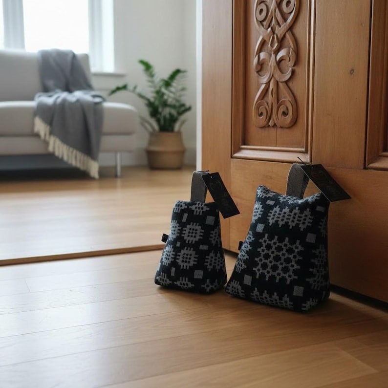 Puede incluir: Dos topes de puerta de tela negra con un patr&oacute;n geom&eacute;trico gris repetitivo. Cada tope de puerta cuenta con un asa de tela gris resistente en la parte superior y una peque&ntilde;a etiqueta rectangular negra unida al lateral, sobre un suelo de madera clara.