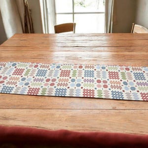 Puede incluir: Un camino de mesa colorido con un patrón geométrico en tonos de azul, rojo, verde y blanco. El camino de mesa está sobre una mesa de madera con dos sillas en el fondo.
