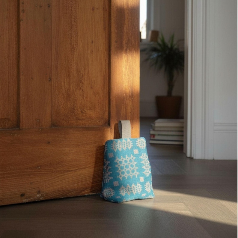 Puede incluir: Un tope de puerta en forma de cu&ntilde;a hecho de tela azul brillante con un patr&oacute;n geom&eacute;trico blanco repetitivo. El tope de puerta tiene un asa de tela gris claro unida a la parte superior, dise&ntilde;ada para mantener una puerta abierta contra una superficie.