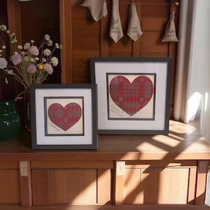 May include: Two framed artworks with a heart design. The hearts are red with a gray pattern, set against a cream background, and framed in dark gray. One artwork has the word "Cwtsh" and the other has "Calon Y Cartref" written on it.