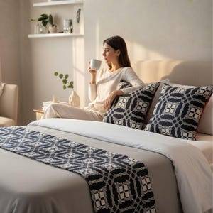 May include: A bedroom scene featuring a bed with a gray comforter, white sheets, and decorative pillows. The pillows and bed runner have a geometric black and white pattern. A person is sitting on the bed, holding a white mug.