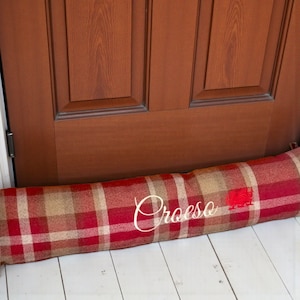 May include: A red, beige, and white plaid door draft stopper with the name "Croese" embroidered in white script. The rectangular pillow is resting on white painted wooden floorboards in front of a brown wooden door.